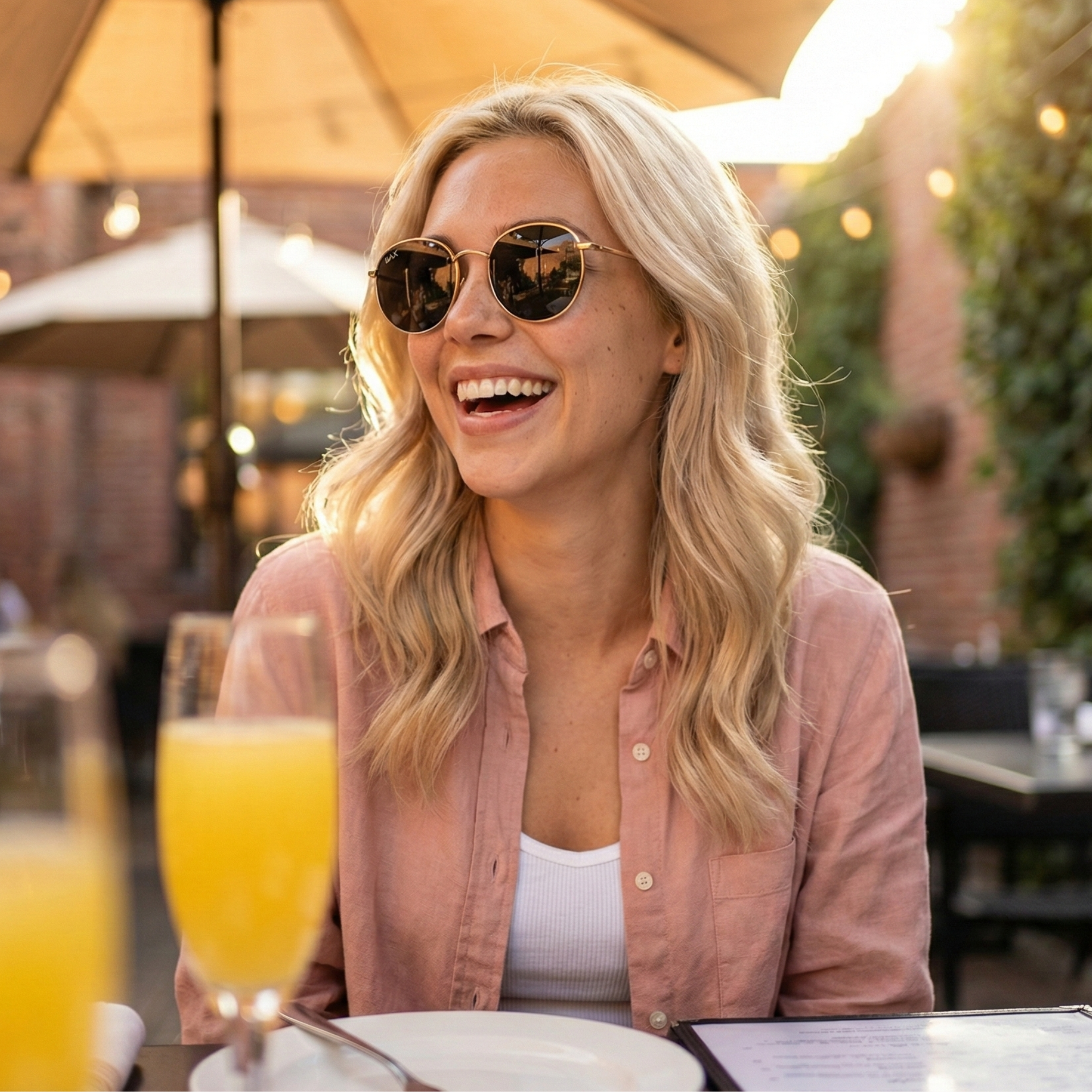 Woman wearing sunglasses and a pink shirt, sitting outdoors with a glass of orange juice.