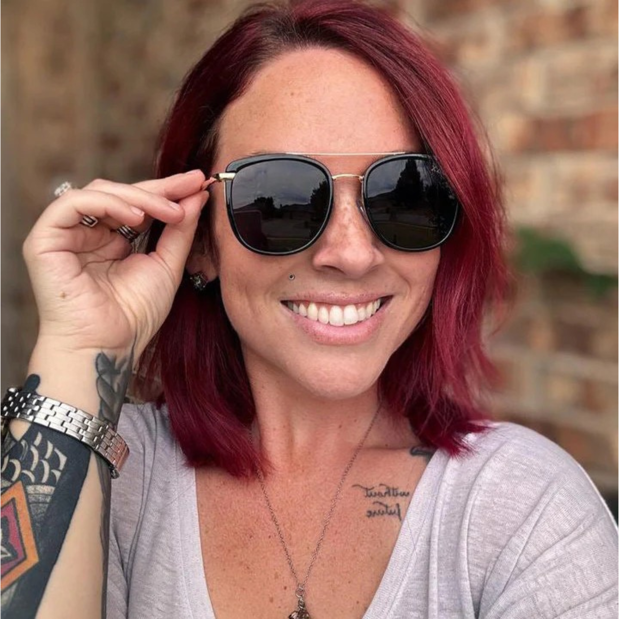 Woman with red hair wearing sunglasses and a gray shirt, standing against a brick wall.