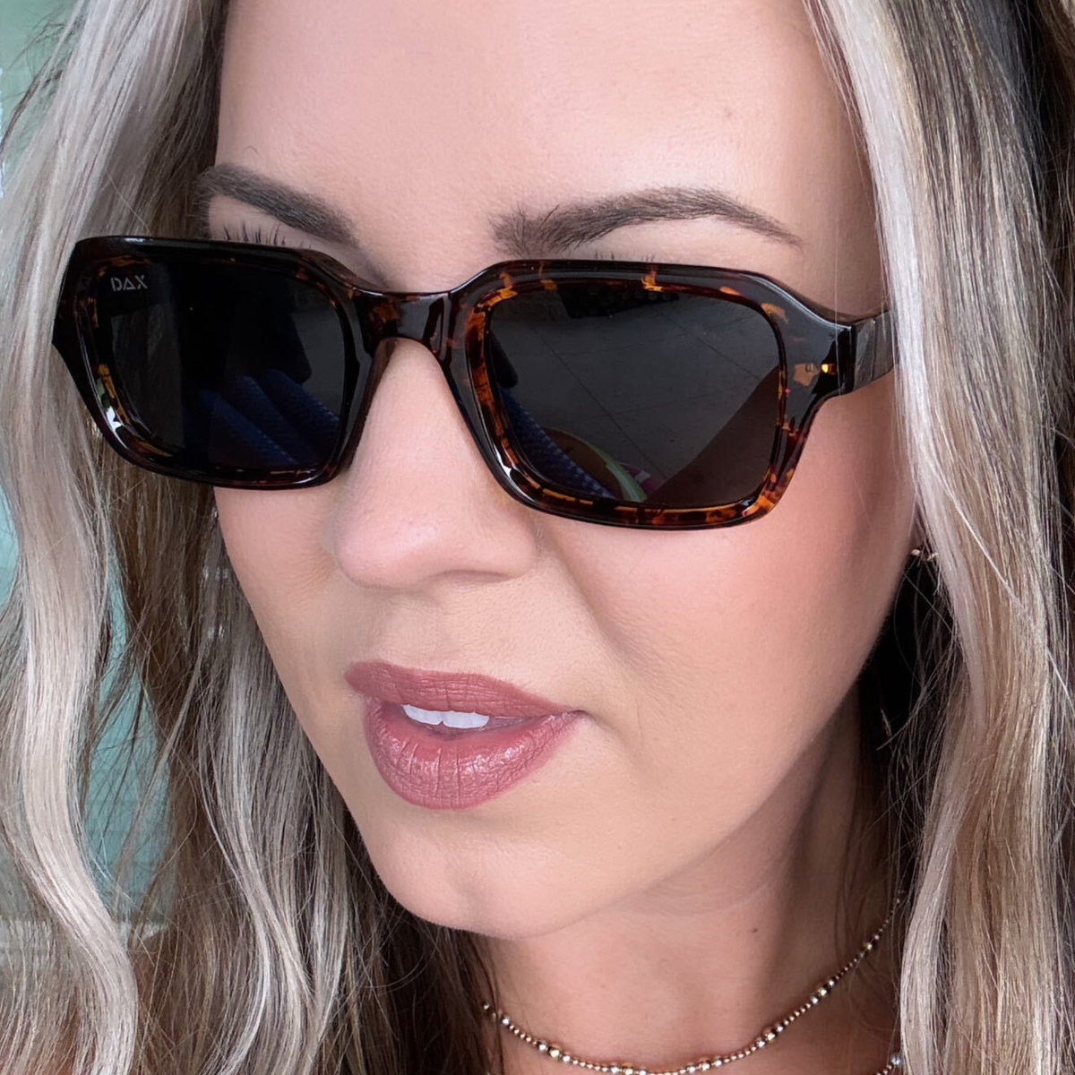 A close-up of a woman wearing dark brown tortoiseshell square sunglasses with dark lenses. The brand name 'dax' is visible on the left lens.