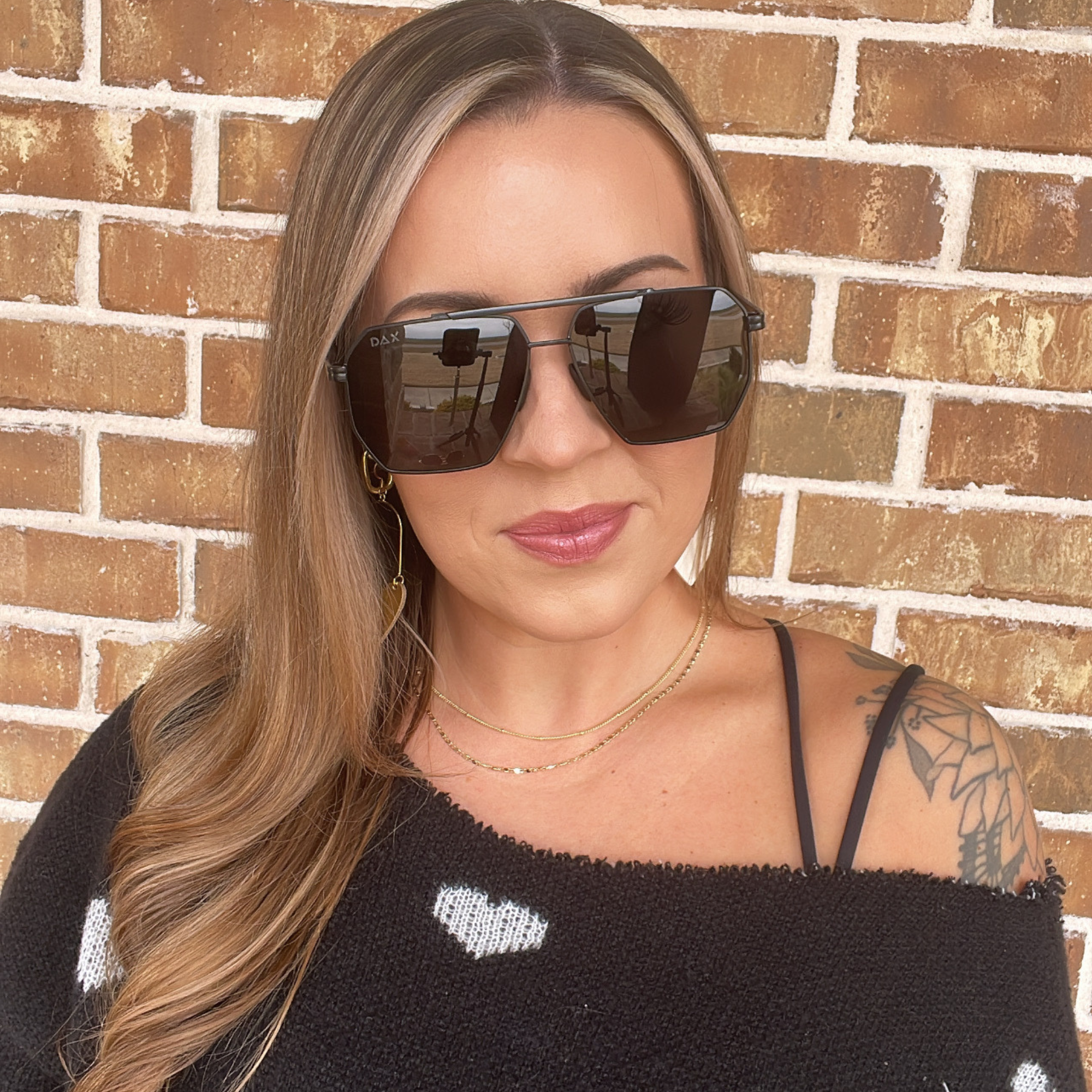 Woman wearing sunglasses and a black top with white patterns against a brick wall.