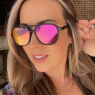 Woman wearing black sunglasses with pink and orange reflective lenses, gold earrings, and necklaces.