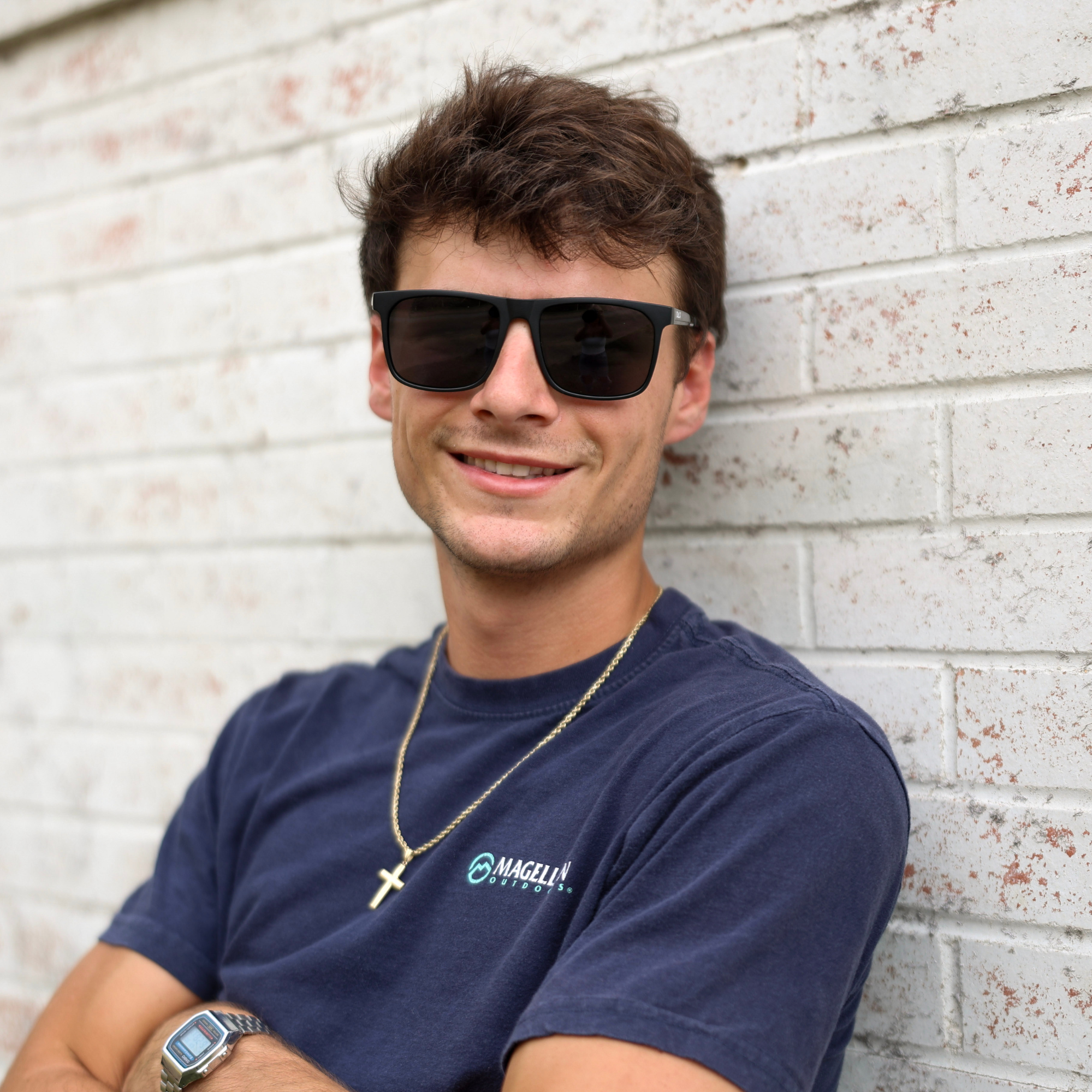 Man wearing sunglasses and a navy blue t-shirt with a logo, leaning against a light-colored brick wall.