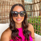 Woman wearing sunglasses and a pink top sitting outdoors with a wicker chair and greenery in the background.