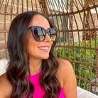 Woman wearing sunglasses and a pink top sitting under a wicker canopy with greenery in the background