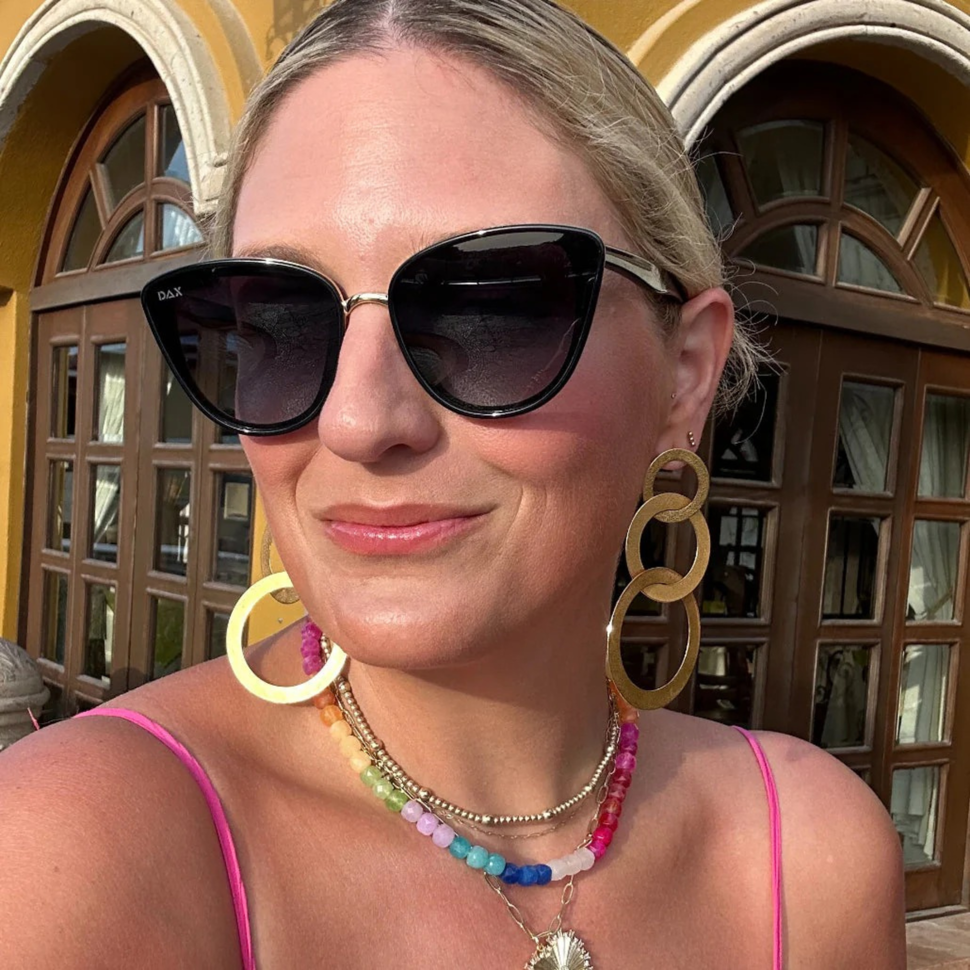 Woman wearing sunglasses and colorful jewelry in front of a building with arched windows.