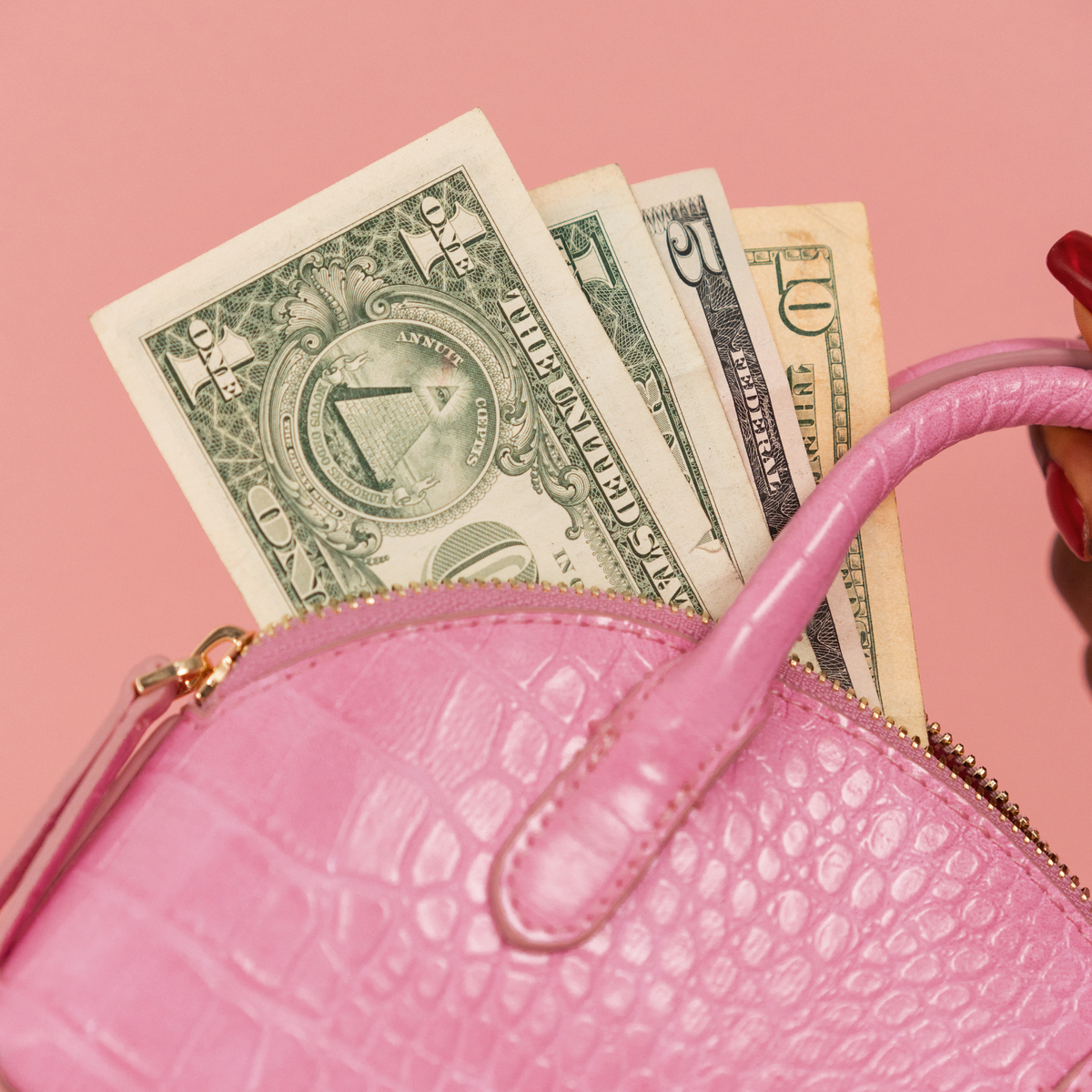 A close-up of a pink crocodile-embossed handbag with a gold zipper, partially filled with U.S.