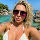 Woman wearing sunglasses and a white bikini top with a scenic background of rocks and water.