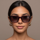 A close-up portrait of a woman wearing oversized square sunglasses with dark brown tinted lenses and dark burgundy frames, paired with gold hoop earrings against a solid taupe background.
